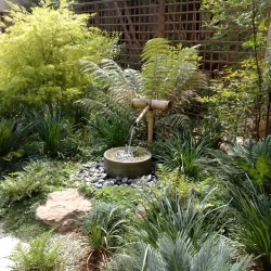 Japanese water basin (tsukubai) with bamboo water feature (kakehi), black pebbles, lush planting.
