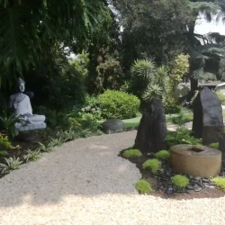 Three monolith rocks and Japanese water basin (tsukubai) set into mossy mound, surrounded by beige gravel in centre of meditation garden. Small curved Zen benches. Large white Buddha ornament.