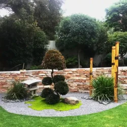 Rounded garden bed. Japanese garden. Low solid granite Japanese lantern. Multi-head shaped cloud tree with mosses. Liriope Green Giant. Pebbles. Gravel. Three tall bamboo poles. Rocks.