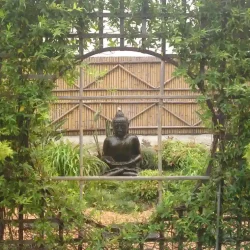 Life-size black Buddha statue surrounded by lush planting and backed by a bamboo wall screen, viewed through a moon gate trellis.