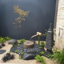 Traditional garden with a modern feel. Japanese water basin (tsukubai) with bamboo kakehi water feature and stone tower. Dark grey rocks and black slate stepping stones, black pebbles, ferns liriope, Black Dragon, and a Japanese Maple tree against the dark grey wall. All surrounded by pale beige gravel.