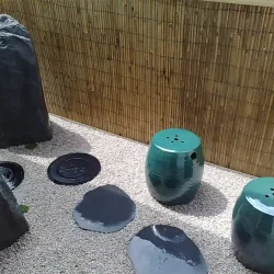 Tiny balcony garden. Path of decorative black round steppingstones, set into gravel leading to a small self-contained Zen water feature. Three monolit rocks, a small potted nandina, very few mossy ground covers and split bamboo cladding to add a finishing touch and texture. 2 tiny ceramic seats/tables