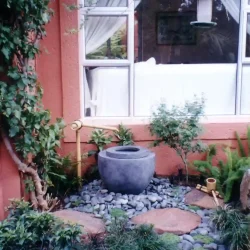 Japanese garden with hand-crafted ceramic Japanese water basin (tsukubai) with bamboo water feature and bamboo ladle & rest, surrounded by black pebbles.