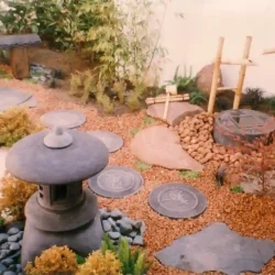 Courtyard garden with Japanese water pot, shishi-odoshi deer scarer water feature, kakehi bamboo water feature, bamboo ladle & rest, round black Japanese lantern, simple rustic wooden bench, decorate Japanese stepping stones, brown gravel.